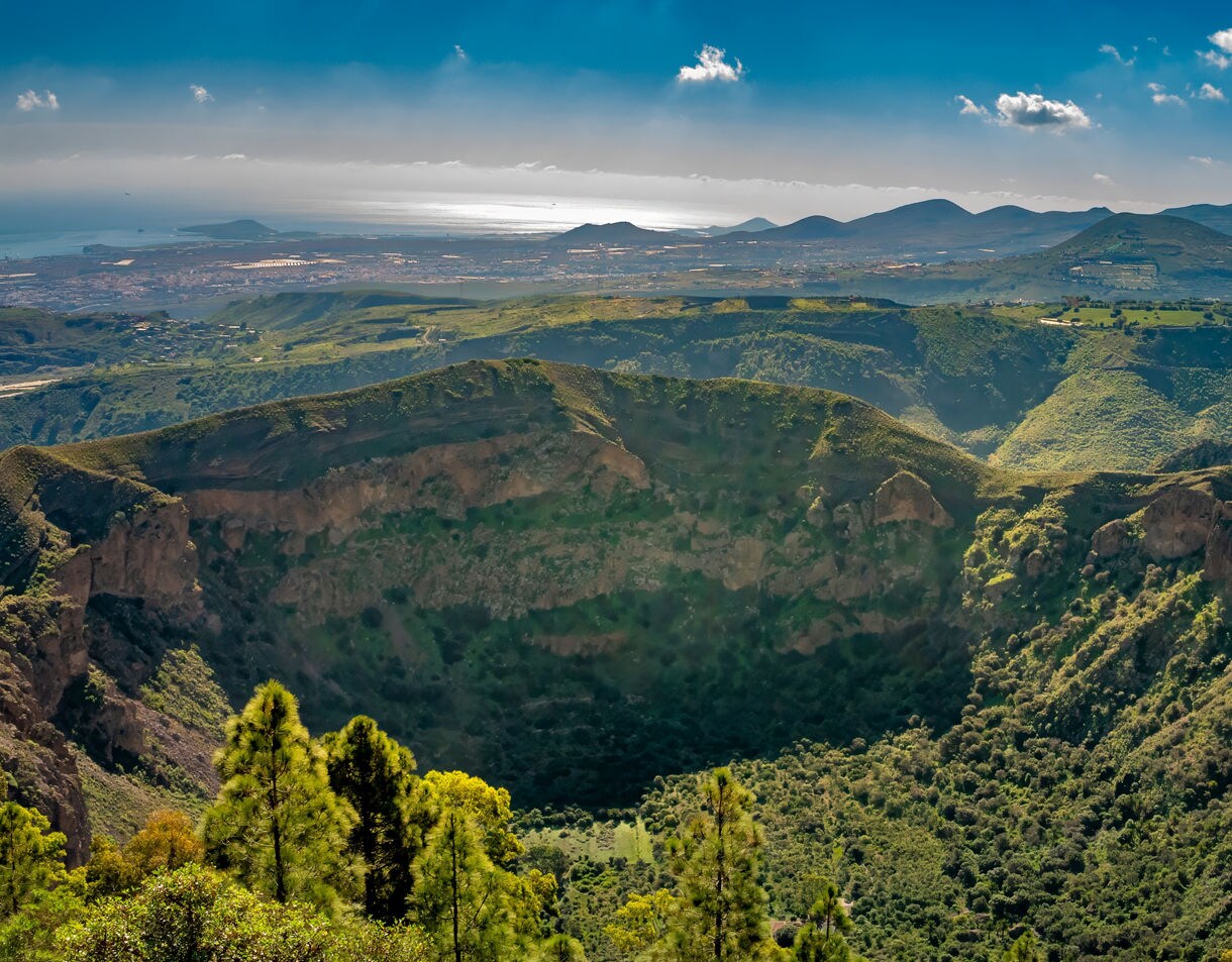Panoramic view of the Bandama Caldera in Gran Canaria, with steep rocky walls, dense vegetation inside the crater and rolling hills stretching toward the coastline.